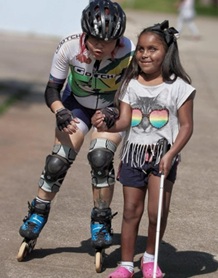 mulher ajoelhada de patins com criança aparentando cerca de 5 anos, segurando uma pequena bengala cor de rosa.